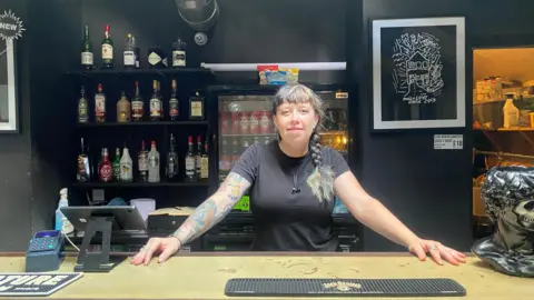 A woman with arm tattoos and braided hair in black T-shirt stands behind a bar. 
