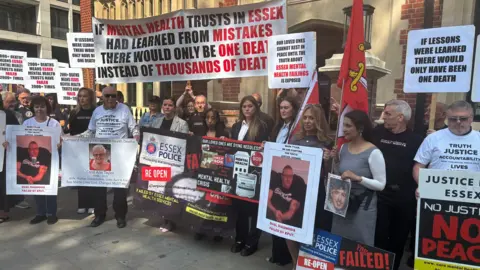 Bereaved families gathering outside the Lampard Inquiry at Arundel House, London. They are holding a variety of banners and photographs of their loved ones who they have lost. The slogans include sentences such as "if lessons were learned there would only have been one death". Some of the family members are wearing white T-shirts with "truth, justice, accountability" written on them.