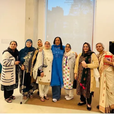 Anjum Malik A group of Asian women in traditional dress stand together in a line in a white room.
