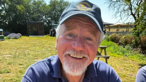 A man in a navy polo shirt and cap smiles in a field. He has a white beard and white hair. Just his head and shoulders can be seen.