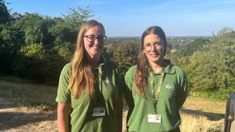 Cass and Megarrell smiling at the camera, both wearing green T-Shirts and Richmond Park passes around their necks. Cass has long blonde and Megarrell long brown hair. They are standing on dried out grass with bushes and blue skyes in the background.