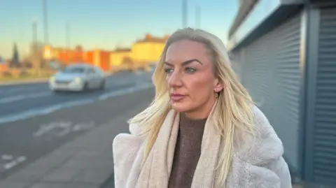 Amy, who has long, blonde hair, looks to her authority. She is wearing a fluffy jacket and it is a sunny afternoon. She is standing in front of closed roller blinds at a shop. A blurred car on a road is in the background.