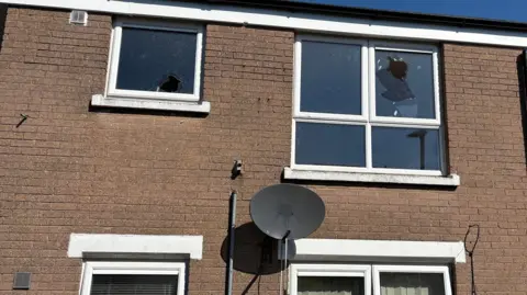 A close-up of the attacked house in Kincraig Avenue, Belfast.  Two of the upstairs windows have been smashed in the two-story, brown brick terraced property.  There is a black satellite dish on the wall and a cloudless blue sky in the background. 
