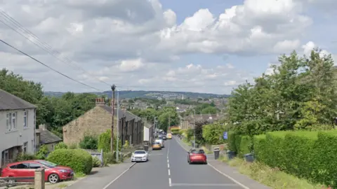 Google A residential street overlooking hills with cars parked alongside hedges, trees and houses.