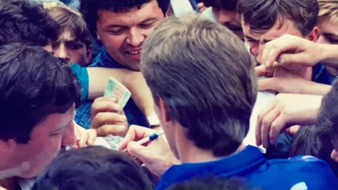 Tony Jones Sergei Baltacha is pictured from behind signing autographs for a busy crowd os people. He has thick hair swept back and is wearing a blue top.