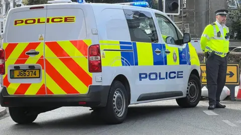 BBC A police van is parked on a road with its blue lights flashing. A police officer in uniform is standing in front of it with his hands held behind his back in front of a road diversion sign.
