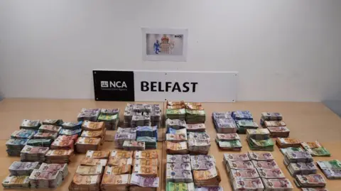 Multiple stacks of bank notes, in brown, blue and green, on display in front of a black and white sign saying NCA Belfast.