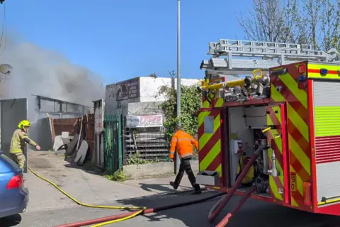 A fire engine and two firefighters are in the foreground. Smoke is billowing from the fire in the background.