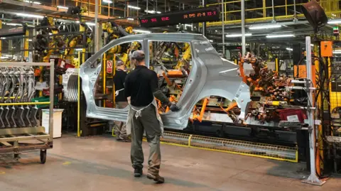 Two workers at Nissan's factory in Sunderland. One man is carrying a large side panel of a car. In the background are machines and robotic equipment used on the assembly lines.