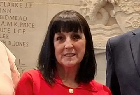 A close up of Lynn Worrall  with dark shoulder-length bobbed hair and a heavy fringe. She is smiling at the camera and wearing a red top with a gold button
