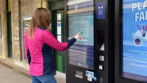 A northern woman in a pink jacket buys a ticket from a northern ticket machine.