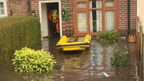 Polly Pee A yellow inflatable raft floats on brown muddy floodwater in the submerged back garden of a house