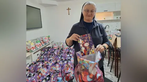 Belveder A nun in a blue and white wimple holds a Cadbury selection box as she puts it in a gift bag. Hundreds of Christmas bags are on the floor behind her and a crucifix is on the wall.