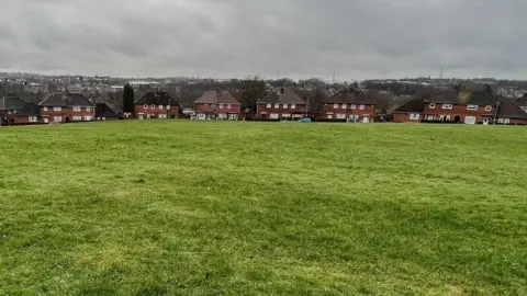 BBC A row of semi-detached houses can be seen with a large open space in front of them.