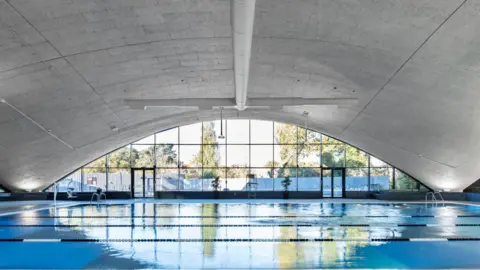 Historic England Archive An interior image of the swimming pol, with grey concrete arching over the pool and a square-panelled arched window at the end.