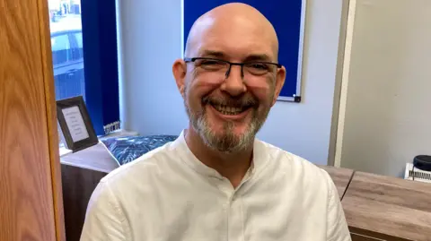 Samuel Goss. He is bald but has grey facial hair. He is wearing glasses and a white shirt. He is looking directly at the camera and smiling. 