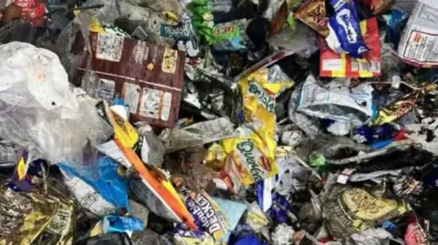 BEACH BUDDIES A close-up of rubbish, which includes multicoloured crisp packets and sweet wrappers.