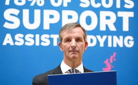 Getty Images Liam McArthur at the launch of his bill in March 2024. He is standing behind a podium and in front of a blue background with the words 78% of Scots support assisted dying.