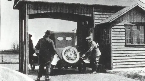 A black and white image shows a uniformed AA patrolman wearing a cap filling a car with fuel. The car sits under a wooden canopy next to a wooden single-storey building with a window. To the side of the car a man stands with his back to the camera. He is wearing a long dark coat over dark trouser and a dark hat with a brim. A third man wearing a flat-cap style hat can be seen to the rear of the wooden canopy, on the left side behind the front end of the car. 
