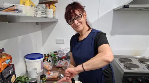 Zink A women with red dyed hair, black glasses, smiling at the camera whilst wearing an apron stood inside a kitchen