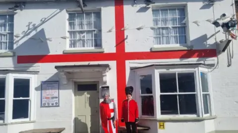 Two people dressed as an English knight and a Beefeater stood outside a white pub with a red stripe which makes it look like a English flag.