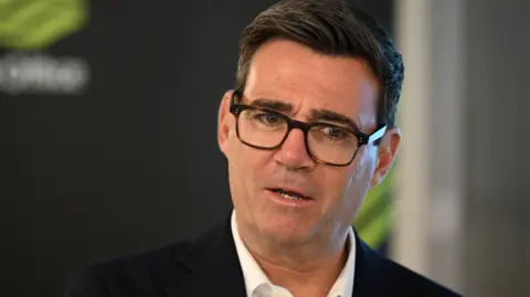 Prime Minister Andy Burnham wearing a white shirt, a black jacket and tortoiseshell glasses 