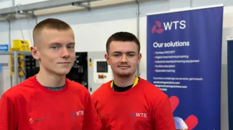 Oli and Joey have very short hair and are in red t-shirts with the logo WTS and are standing inside the Energy Skill Learning Hub. There is a purple sign behind them saying WTS - Our solutions but the rest of the writing is out of focus.