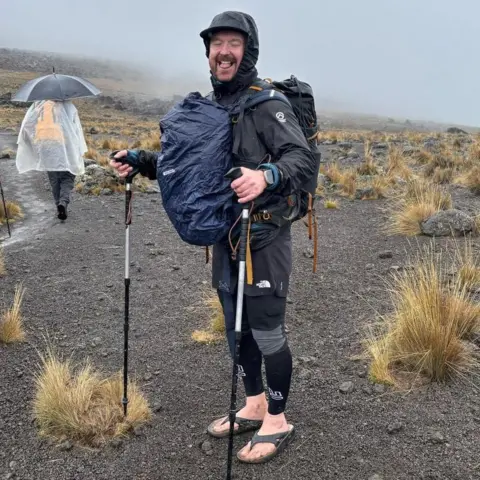 Stu Skinner A laughing Stu Skinner pokes his tongue out for the camera during a rainy hike. He is wearing black hiking gear with a hood and carrying two full backpacks on his front and back. He is also holding Nordic hiking poles in his hands. His feet are bare with the exception of a pair of flip flops. The landscape is bare dark grey stone, dotted large spiky tufts of grass. A person in the background is wearing a poncho and carrying an umbrella.