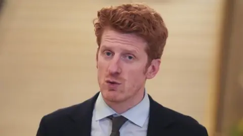 PA Media Matthew O'Toole, a Stormont MLA, speaking in front of the camera. He has red hair and blue eyes and is wearing a dark suit, shirt and tie.