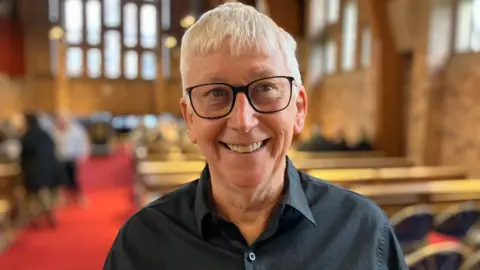 A man with white hair, black framed glasses in a black shirt looking at the camera with a smile
