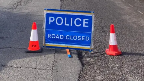 BBC A blue Police Road Closed sign with an orange traffic cone either side on a tarmac road