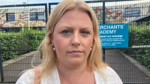 Kerry wearing a white top with blonde hair standing in front of school gates looking into the camera