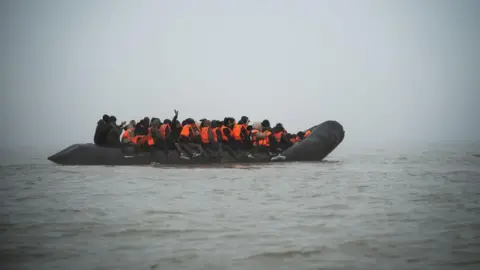 A black rubber dinghy filled with passengers, most wearing orange life vests, floats in fog on the English Channel. One person raises their hand.