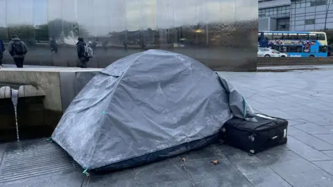A grey tent on the pavement. People in thick coats walk past.