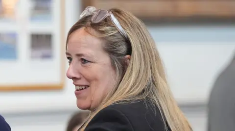 PA Media Susan Davy with her back to the camera and looking to her left with a pair of glasses on her head and smiling in a room during a meeting.