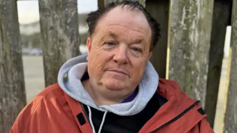 David Moores has black hair and is wearing a grey hoodie under a red and black coat. He is sitting in front of wooden fencing.