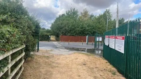 An image of two halves of a grass track, split by a tarmac road. A faded black and white crossing denotes where walkers can cross. The grass is dry and yellow. Hedgerows trees and a green fence border the track. "Warning" signs are attached to the fence.