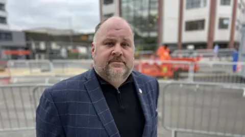 Leader of Swindon Borough Council, Jim Robbins, wearing a navy blue check jacket standing in front of the construction site