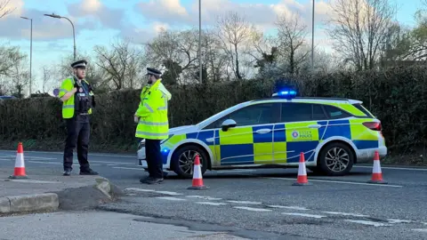 Stuart Woodward/BBC A police car is parked across a road with two police officers stood next to it. Orange cones block the road