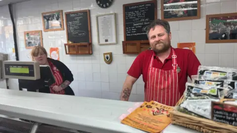 Jonny Humphries/BBC Danny Shaw, in a red polo-neck t-shirt with a red and white butcher's apron, stands behind the counter of his butcher's shop. Another member of staff, a woman in a black cardigan and also wearing an apron, is to the left