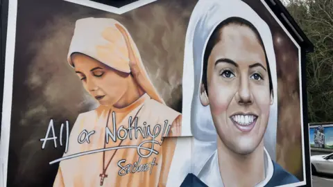 Shows a mural with two images of the nun Sr Clare Crockett. On the left, she is wearing a light orange habit and veil, her head bowed and a cross around her neck. The words All or Nothing!! are written over it, with Dr Claire underneath. On the right she is wearing a white veil and blue and white habit and smiling. 