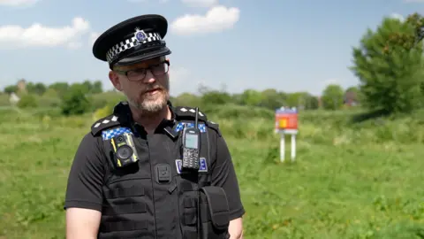A police officer in black uniform with greenery behind him, including a trip and a sign