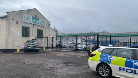 A large light brown building with a sloped roof and metal railings at the front and a police car parked in the foreground