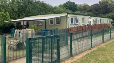 Green portable cabin with windows and a white door. There is a brown wooden framed-fence around it with a green play area in front of it. There is a green fence separating the pre-school from a walkway. 