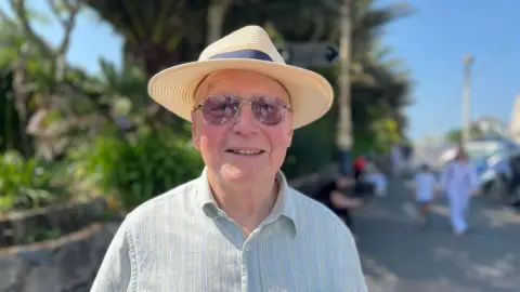George Carden / BBC A man, Peter Austin, wearing a light coloured shirt, a hat and sunglasses. He is stood in a street.