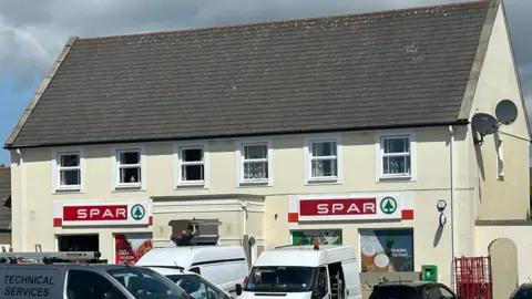 The exterior of the Spar, a cream building with a grey slanted roof, signs at either side of the entrance read Spar.