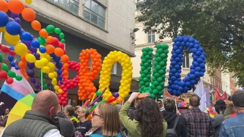A rainbow collection of balloons spell out "proud" above the heads of a crowd of people on a street.