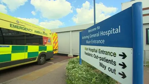 Entrance to Kettering General Hospital showing blue and white Welcome sign and yellow and green ambulance with rear doors open