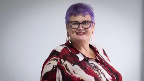Getty Images Carolyn Harris MP poses for a photograph. She has purple hair, has glasses on and is wearing a red and white top.