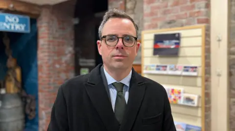 A man wearing a black coat over a suit with a blue shirt and grey tie, wearing black glasses standing in a museum - the background is blurred.